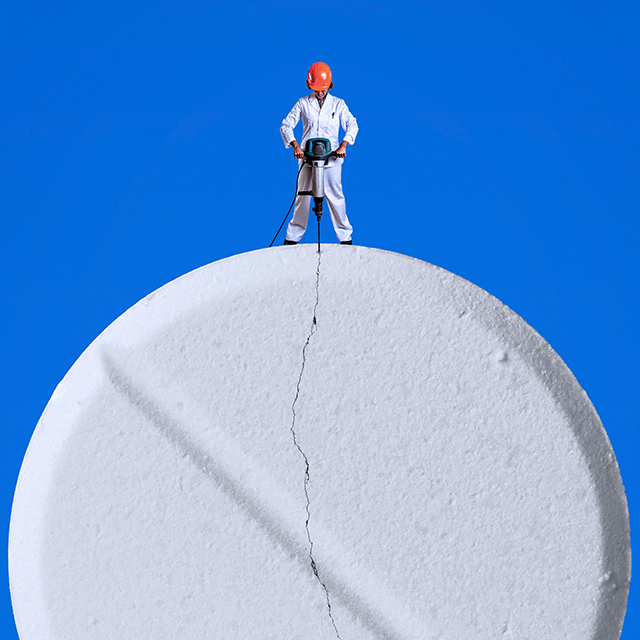 A woman wearing a white uniform and a red hard hat jackhammers a cracked, giant pill.