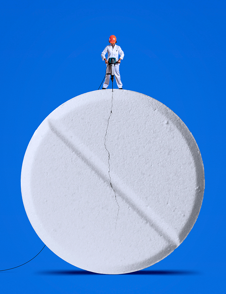 A woman wearing a white uniform and a red hard hat jackhammers a cracked, giant pill.
