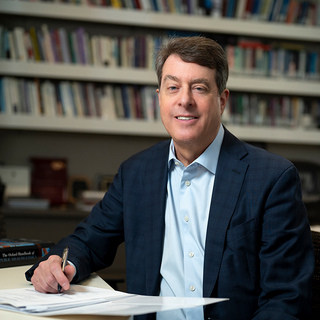 Jeffrey Khan sits at a desk, pen in hand, as he smiles at the camera.