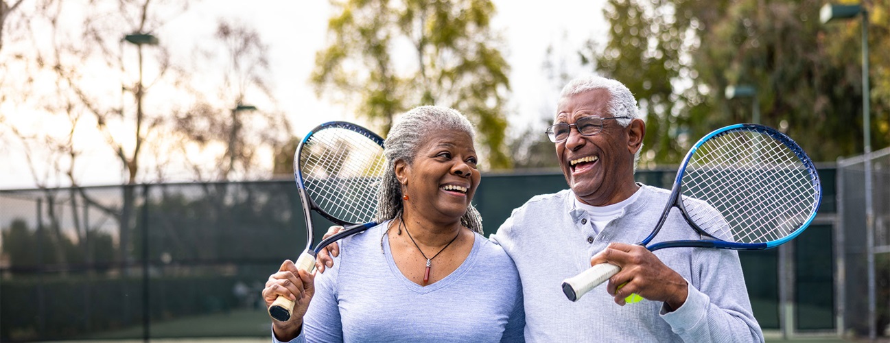 A older  man and woman couple on a tennis course with rackets in their hands. The man has his arm around the woman, they are both laughing.