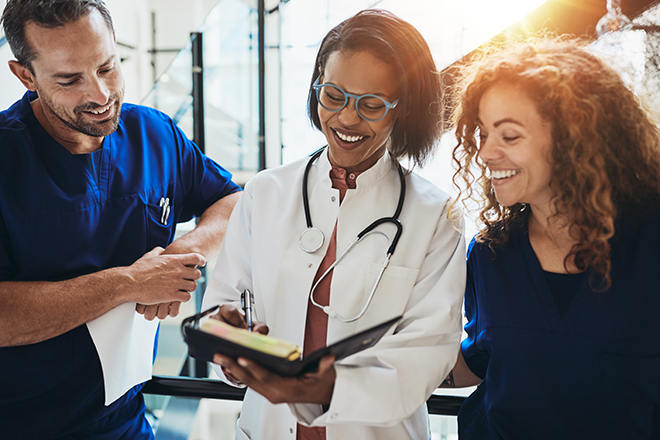 An smiling, diverse team of three healthcare professionals in scrubs and a white lab coat stand together, looking at a notepad while reviewing medical information.