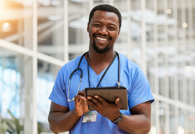 Smiling doctor with stethescope around his neck, in blue scrubs holding a tablet device.