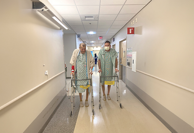 Two men in hospital gowns walking down a hospital hallway.