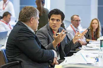 Men in suits sitting at a table covered in paper, discussing something seriously