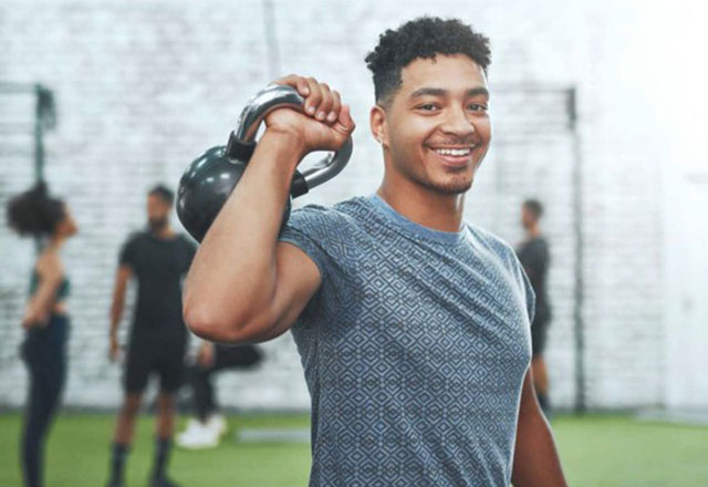 A man standing in a gym holding a kettlebell over his shoulder.