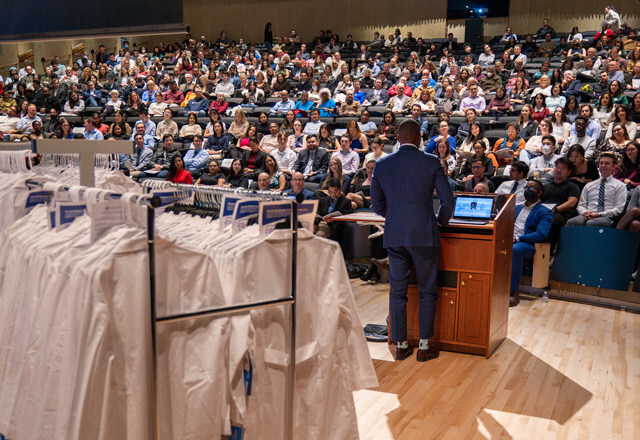 View from back of stage featuring racks of white coats, speaker at podium, and full audience.