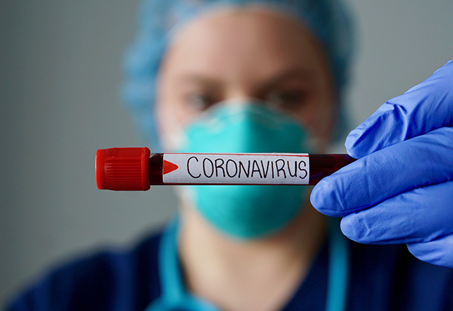 A healthcare worker wearing a mask holds up a test tube labeled, &quot;Coronavirus.&quot;