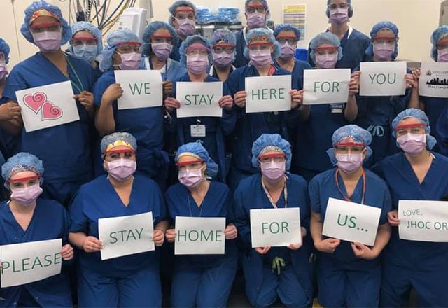 Hopkins staff, dressed in scrubs and masks, hold up signs that read 'We stay here for you. Please stay home for us... Love, JHOC OR.'